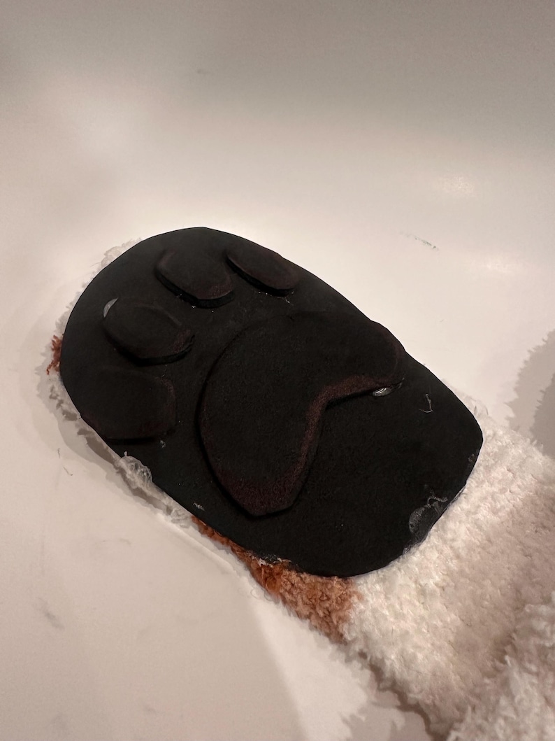May include: A black dog paw-shaped item with three oval pads and a larger pad, set against a white and brown fluffy background. The paw is made of a dark, possibly felt-like material.