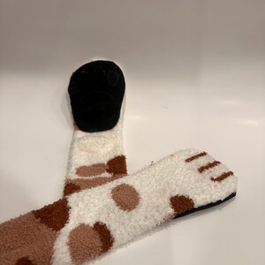 May include: Cozy, white and brown patterned socks with a black paw-shaped design. The socks have a soft, fuzzy texture and a playful, animal-inspired design, perfect for lounging or adding a fun touch to any outfit.