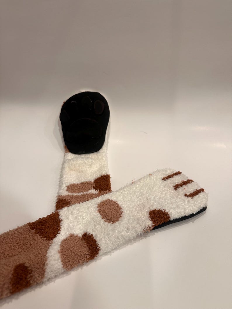 May include: Cozy, white and brown patterned socks with a black paw-shaped design. The socks have a soft, fuzzy texture and a playful, animal-inspired design, perfect for lounging or adding a fun touch to any outfit.