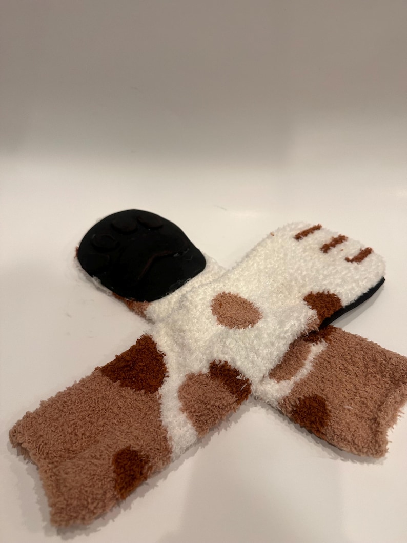 May include: A pair of cozy socks with a paw print design. The socks are white with brown spots and a brown cuff. The soles are black.