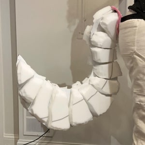 May include: A curved, segmented tail prop constructed from white foam blocks. The tail features a series of stacked, rounded foam pieces that create a flexible, articulated appearance. A small pink accent is visible at the base attachment point.