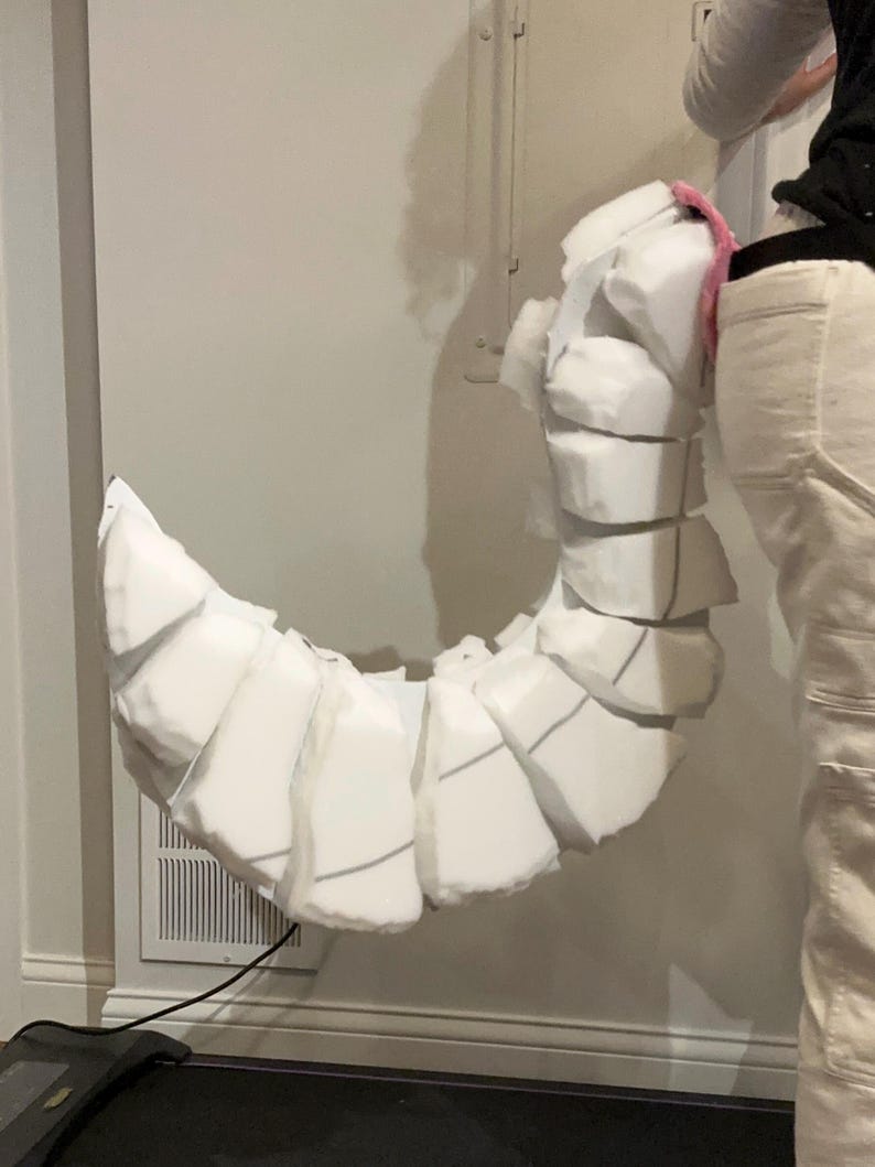 May include: A curved, segmented tail prop constructed from white foam blocks. The tail features a series of stacked, rounded foam pieces that create a flexible, articulated appearance. A small pink accent is visible at the base attachment point.