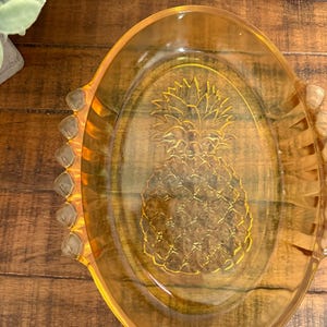 May include: Amber-coloured glass serving dish with an oval shape and a pineapple design in the centre. The dish has two handles and a textured rim. The pineapple design is etched into the glass.