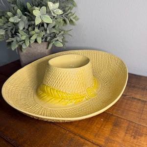 Könnte beinhalten: Eine gelbe, keramik Sombrero-förmige Servierschale mit strukturiertem Design. Die Schale hat eine zentrale Schüssel und einen breiten Rand, mit einem dekorativen Blatt- und Wirbeldesign am Hutrand. Die Schale steht auf einer Holzoberfläche.