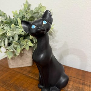 以下が含まれることがあります： 鮮やかな青い目の黒いセラミック製の猫の置物。猫は直立して座っており、耳を立てています。置物は木製の表面に置かれ、背景には鉢植えの植物があります。