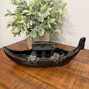 Op de afbeelding: Een zwarte keramische gondelvormige plantenbak met een kleine cabine en bloemaccenten. De plantenbak staat op een houten oppervlak, met een groene plant in een pot op de achtergrond. De gondel is ongeveer 30 cm lang.