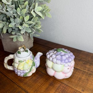 Op de afbeelding: Een keramische theepot en suikerpot set met een fruitontwerp. De theepot heeft een gouden handvat en deksel. De suikerpot heeft een paars druivenontwerp.