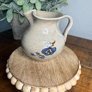 May include: A small, beige ceramic pitcher with a handle and a spout. It features a blue floral design and sits on a round, wooden beaded stand. The pitcher is a decorative item.