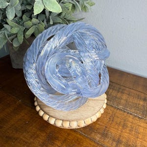 May include: A decorative glass knot sculpture in shades of blue and white, resting on a round wooden base with a beaded edge. The sculpture is displayed on a wooden table, with a potted plant in the background.