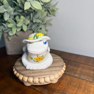 May include: A white ceramic frog ornament wearing a hat with yellow flower accents and a yellow flower design on its chest. The frog is on a small, round wooden stand with a beaded edge.