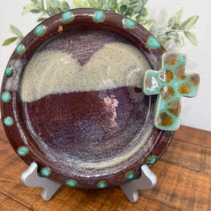 Könnte beinhalten: Ein handgefertigter Keramikteller mit Kreuzakzent. Der Teller hat eine tief burgunderrote Farbe mit einem beige-violetten Innenraum. Türkisfarbene Punkte zieren den Rand, und ein türkisfarbenes und braunes Kreuz ist an der Seite befestigt. Der Teller wird auf einem silbernen Ständer präsentiert.