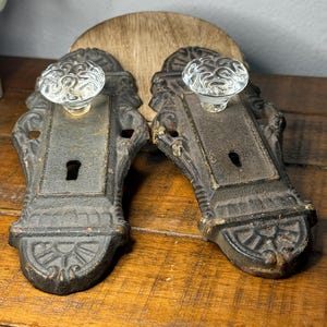 May include: Two matching door plates made of dark, weathered cast iron, each featuring an ornate, rectangular design with a central keyhole and a clear, faceted glass knob attached to the top section of the metal hardware.