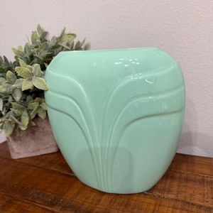 Könnte beinhalten: Hellgrüne Keramikvase mit abgerundeter Form und einem dekorativen, stilisierten Blattdesign. Die Vase steht auf einer Holzoberfläche, im Hintergrund ist eine kleine Topfpflanze zu sehen.