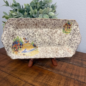Op de afbeelding: Een rechthoekige serveerschaal in vintage-stijl met een crèmekleurige achtergrond en een gouden bloemenpatroon. De schaal heeft twee geschilderde scènes van een vrouw in een jurk en een bloemstuk. De schaal wordt op een houten standaard gepresenteerd.