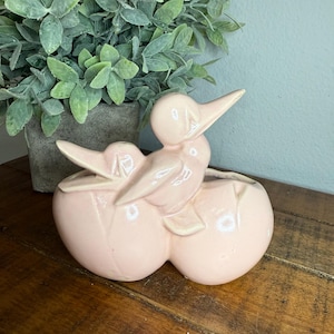 May include: A pink ceramic duck figurine with two ducklings emerging from eggs. The figurine is glossy and sits on a wooden surface. A green plant in a gray pot is in the background.