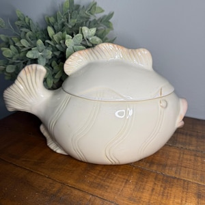 Peut inclure: Un récipient en céramique en forme de poisson avec un couvercle. Le récipient est blanc cassé avec des accents roses sur les nageoires et la bouche. Le poisson a des lignes gravées et un œil rond. Le récipient est sur une surface en bois.