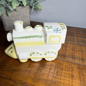 May include: A ceramic train figurine in white, yellow, and green. The train features a locomotive, a passenger car, and decorative floral accents. The figurine is placed on a wooden surface.