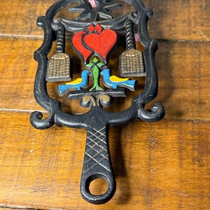 Puede incluir: Un salvamanteles de hierro fundido negro con un corazón rojo central, dos pájaros azules y amarillos y dos formas de escoba de color beige. El marco tiene una estrella en la parte superior y un mango con textura de rombos y un agujero.