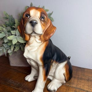 May include: A ceramic figurine of a beagle dog in a sitting position. The dog features a tri-color coat with patches of black, brown, and white. It has large, dark, expressive eyes, a black nose, and long, drooping ears with a smooth finish.