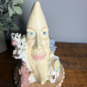 May include: A whimsical, cream-colored moon-shaped figurine with blue eyes and a smiling face. The figurine features a cow, pig, and cat, and is set on a wooden base. A unique decorative piece.