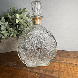 Può includere: Un'antica caraffa di vetro trasparente con un motivo decorativo a stella. La caraffa ha un corpo rotondo, una base quadrata e un tappo di vetro trasparente. Un tappo di sughero sigilla la bottiglia. La caraffa è su una superficie di legno.
