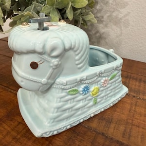 Op de afbeelding: Een lichtblauwe keramische schommelpaardplanter met een mandweefontwerp. Het paard heeft een bruin oog en een hoofdstel. De planter is versierd met kleurrijke bloemen en is geschikt voor kleine planten of bloemen.