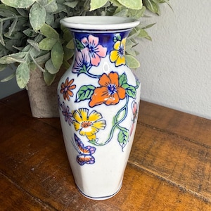 Può includere: Un vaso di ceramica bianco con bordo blu e design floreale. Il vaso presenta fiori arancioni, gialli e rosa, insieme a foglie verdi e una farfalla. Il vaso è alto circa 15 cm.