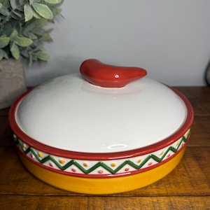 May include: A ceramic serving dish with a lid, featuring a white top and a yellow base. The rim is decorated with a red border and a green, yellow, and orange zigzag pattern. A red chili pepper serves as the handle.