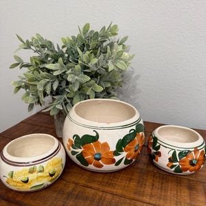 May include: Three ceramic bowls with floral designs. The largest bowl features orange flowers with blue centers and green leaves. The other two bowls have similar designs, with one featuring yellow flowers. The bowls are on a wooden surface.