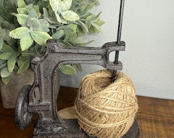 May include: A dark gray cast iron twine holder featuring a vintage sewing machine frame design with a vertical rod for holding a ball of natural tan jute twine. The heavy metal base supports the frame and the twine, which is wound into a sphere.