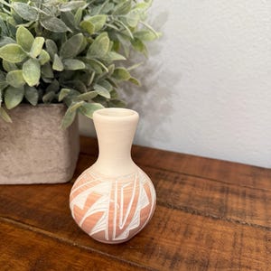 May include: A small, off-white ceramic vase with a rounded body and a narrow neck. The vase features a peach-colored base with a white geometric design. The vase is displayed on a wooden surface, with a potted plant in the background.