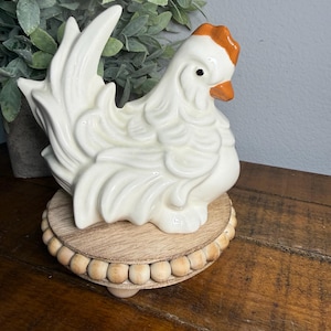 Puede incluir: Figura de gallina de cerámica blanca con detalles en naranja en la cresta y el pico, colocada sobre una base redonda de madera con borde de cuentas. La gallina presenta plumas detalladas y un acabado brillante, ideal para decoración rústica.
