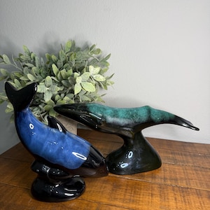 Puede incluir: Dos figuras de ballenas de cerámica. Una es azul y negra, la otra verde y negra. Las figuras están sobre una superficie de madera con una planta verde en el fondo. Las ballenas tienen un acabado brillante.