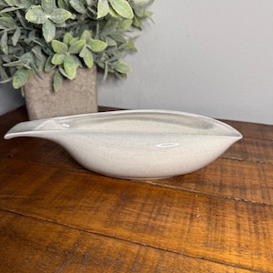 Puede incluir: Un cuenco de cerámica oblongo gris claro con un acabado moteado. El cuenco tiene una forma única y alargada con bordes ligeramente elevados. El cuenco es probablemente para fines decorativos o de servicio. Una planta verde en una maceta cuadrada está en el fondo.