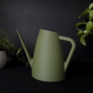 May include: A sage green watering can with a long spout and a curved handle. The watering can has a conical body and is made of a solid material. The background is dark, with some greenery visible on the right and left.