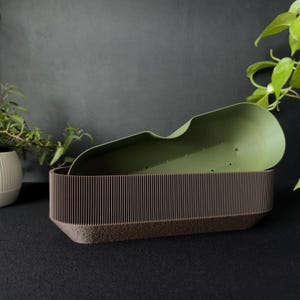 May include: A two-toned planter with a brown base and a ribbed, dark brown upper section. A green, scoop-shaped insert rests inside. Green foliage is visible in the background, creating a natural aesthetic. The planter is designed for indoor or outdoor use.