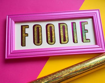 Pode incluir: Uma moldura retangular rosa contendo uma placa branca com a palavra FOODIE em letras grandes, tridimensionais e com glitter dourado. Abaixo da moldura, há um rolo de material com glitter dourado e superfície cintilante.