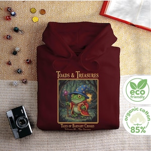 Op de afbeelding: Een bordeauxrode hoodie met de tekst "TOADS & TREASURES" en een illustratie van een kikker-tovenaar met een blauwe hoed en rode mantel. De hoodie heeft ook een "eco friendly" en "organic cotton 85%" label.