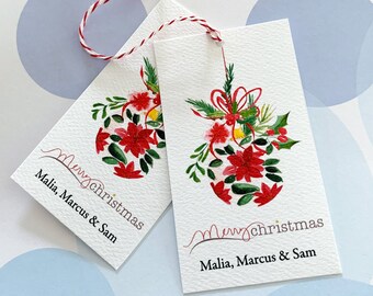 May include: Two white Christmas gift tags with red and green floral designs. The tags feature the words "Merry Christmas" and the names "Malia, Marcus & Sam". Each tag is tied with a red and white twine.