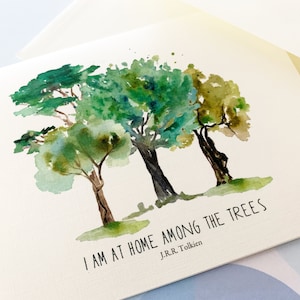 May include: Watercolor art print featuring three trees in shades of green and brown. The text "I AM AT HOME AMONG THE TREES" and the author's name, J.R.R. Tolkien, are printed below the trees. The print is on a cream-colored card.