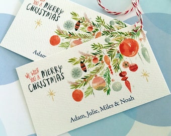 May include: Two rectangular Christmas gift tags with watercolor illustrations of ornaments and greenery. The tags read "WE WISH YOU A MERRY CHRISTMAS" and include names. A red and white string is attached to the tags.