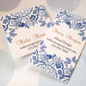 May include: Two business cards with a floral design. The cards are white with a blue floral border and the name "Malia Morgan" in gold script. The cards also include contact information.