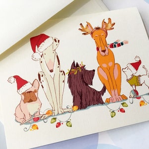 May include: A Christmas card featuring five cartoon dogs. A Dalmatian, a French Bulldog, a Scottish Terrier, a Great Dane, and a small white dog are wearing Santa hats and festive accessories. They are sitting on a string of Christmas lights.