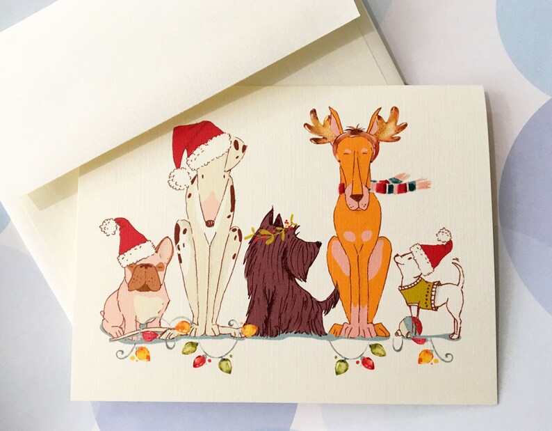 May include: A greeting card featuring a whimsical illustration of five dogs dressed in festive holiday attire. The dogs are adorned with Santa hats, antlers, and a scarf, sitting on a string of lights. The card is accompanied by a cream-colored envelope.