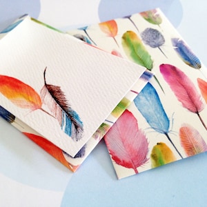 May include: Two folded stationery cards with watercolor feather designs. One card features two feathers in shades of red, orange, and blue. The other card has a pattern of colorful feathers in pink, blue, green, and yellow. The cards are on a light blue surface.