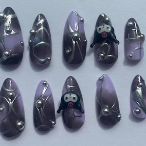 May include: Set of ten lavender and brown press-on nails. Each nail features silver accents, including lines and small, round embellishments. Some nails have a cartoon character design.