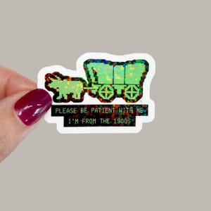 May include: A holographic sticker featuring a pixelated covered wagon and oxen. The sticker has the text "PLEASE BE PATIENT WITH ME I'M FROM THE 1900S" in a pixelated font. The sticker is white with a holographic design.