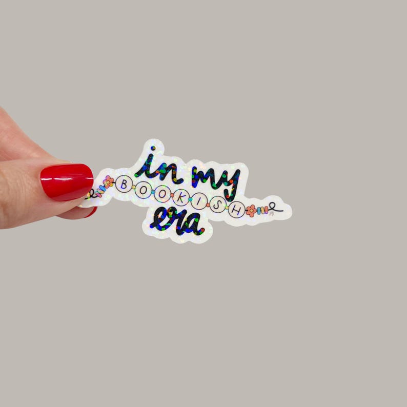 May include: A holographic sticker with the text "in my bookish era" in a whimsical font. The sticker has a white border and is decorated with small floral and circular elements. The background is a solid, neutral gray.