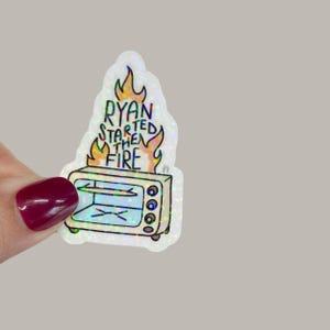 May include: A holographic sticker featuring a cartoon oven engulfed in flames. The text "RYAN STARTED THE FIRE" is written above the oven in a playful font. The sticker has a white border and a glittery finish.