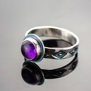 May include: A silver ring with a large, round, purple gemstone. The ring has a blue accent around the gemstone and diamond-shaped patterns etched into the band. The ring is displayed on a reflective surface.